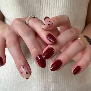 May include: Close-up of hands with oval-shaped nails painted a deep burgundy red. Some nails have a sheer, light pink base with small red hearts.