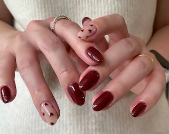 May include: Close-up of hands with oval-shaped nails painted a deep burgundy red. Some nails have a sheer, light pink base with small red hearts.