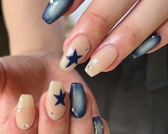 May include: Close-up of a hand with several nails painted in a light beige color with a dark blue star design. Some nails have a gradient effect with a dark blue color fading to light beige.