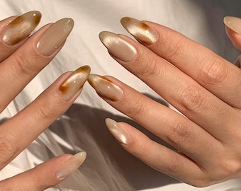 May include: Close-up of a hand with almond-shaped nails painted with a light beige nail polish. Some nails have a brown and gold swirl design.