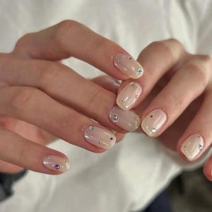 May include: Close-up of a hand with nude oval-shaped nails decorated with small iridescent rhinestones.