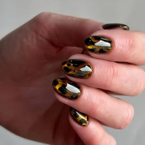 Puede incluir: Uñas ovaladas y brillantes con un diseño de estampado de leopardo en tonos marrón y dorado. Las uñas tienen una superficie pulida y reflectante, creando una manicura elegante.