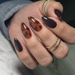Puede incluir: Primer plano de una mano con uñas en forma de almendra pintadas con un patrón de caparazón de tortuga y un esmalte marrón oscuro. Las uñas son largas y puntiagudas.