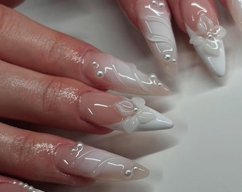 Op de afbeelding: Witte stiletto nagels met een 3D bloemenontwerp en parel accenten. De nagels zijn lang en puntig, met een witte basis en een heldere, iriserende afwerking.