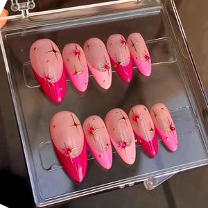 Pode incluir: Um conjunto de dez unhas postiças rosa e vermelhas com glitter dourado e strass. As unhas estão em uma caixa de plástico transparente.