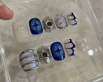Könnte beinhalten: Ein Set mit zehn künstlichen Nägeln mit verschiedenen Designs. Die Nägel befinden sich in einem durchsichtigen Plastikbehälter. Die Designs umfassen ein silbernes geometrisches Muster, einen blauen Stern und einen silbernen und weißen Wirbel.