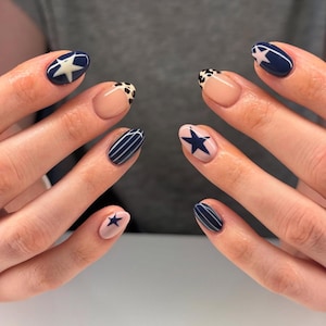 May include: Manicured nails with diverse designs. Some nails are navy blue with white stars, others have white stripes on navy, and some have a nude base with black and white spots or a navy star on a pink base. The nails are oval-shaped.