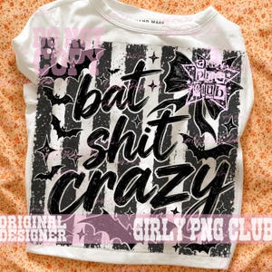 May include: A white t-shirt featuring a black and white vertical striped graphic with the text "bat shit crazy" in a bold, brush-style font. The design includes black bat silhouettes, small four-pointed stars, and a large bow with a star pattern.