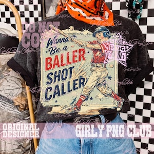 May include: A dark gray t-shirt featuring a baseball-themed graphic. The graphic depicts a baseball player swinging a bat, with the text "Wanna Be a BALLER SHOT CALLER" in a vintage style. The shirt is worn with jeans and an orange bandana.
