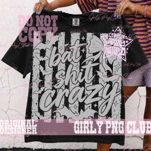 May include: A black t-shirt featuring a large graphic print with vertical stripes, scattered bat silhouettes, and star shapes. The text "bat shit crazy" is printed in a large, stylized, light-colored font across the center of the design.