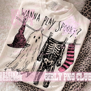 Op de afbeelding: Een cr&egrave;mekleurig t-shirt met een zwarte afbeelding van een heksenhoed, een geest, een skelet en een gestreepte sok aan een waslijn. De tekst "WANNA PLAY SPOOKY?" staat in het zwart boven de items. Het shirt heeft een Comfort Colors label.