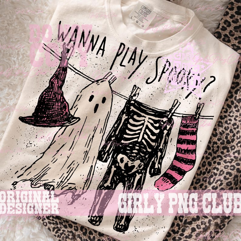 Op de afbeelding: Een cr&egrave;mekleurig t-shirt met een zwarte afbeelding van een heksenhoed, een geest, een skelet en een gestreepte sok aan een waslijn. De tekst "WANNA PLAY SPOOKY?" staat in het zwart boven de items. Het shirt heeft een Comfort Colors label.