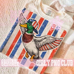 Pu&ograve; includere: T-shirt color crema con grafica di un'anatra con cappello patriottico, con strisce rosse, bianche e blu. L'anatra ha dettagli verdi, marroni e gialli. La maglietta ha il testo "GIRLY PNG CLUB" e "ORIGINAL DESIGNER".