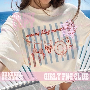 Op de afbeelding: Cr&egrave;mekleurig T-shirt met een strandthema. Het ontwerp omvat een waslijn met een bikini, zwembroek en zonnehoed. De tekst "wanna play merms" staat boven de waslijn.