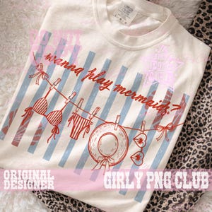 Op de afbeelding: Cr&egrave;mekleurig T-shirt met een rode en blauwe afbeelding. Het ontwerp toont een waslijn met een bikinitop, -broekje, een zonnehoed en hartvormige zonnebrillen. De tekst "wanna play mermaids?" staat erboven.