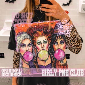 May include: A black t-shirt featuring a graphic illustration of three women with distinct hair colors blowing large bubbles with gum. The design includes vibrant orange, purple, and green tones, with a star-shaped logo containing the text Girly Png Club.