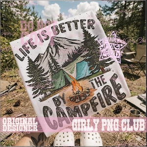 May include: White t-shirt with a camping scene featuring a mountain, trees, tents, and a campfire. The text "LIFE IS BETTER BY THE CAMPFIRE" is printed on the shirt in a bold, stylized font. The design evokes a sense of outdoor adventure.
