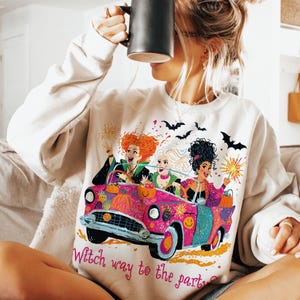 May include: A cream-colored crewneck sweatshirt featuring a graphic print of three characters with distinct hairstyles riding in a bright pink convertible car. The design includes pumpkins, bats, and sparklers, with the text Witch way to the party?