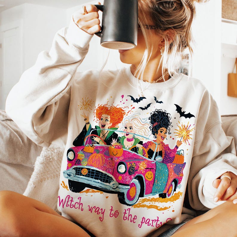 May include: A cream-colored crewneck sweatshirt featuring a graphic print of three characters with distinct hairstyles riding in a bright pink convertible car. The design includes pumpkins, bats, and sparklers, with the text Witch way to the party?