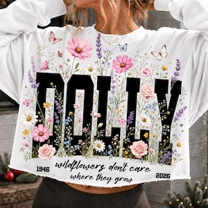 May include: A white long-sleeve sweatshirt featuring the black text DOLLY in a bold font, overlaid with a colorful floral print of pink, yellow, and blue wildflowers, lavender, and butterflies. The text reads 1946 wildflowers don't care where they grow 2026.
