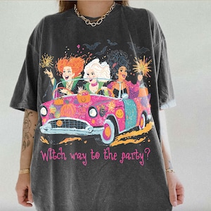May include: A charcoal gray short-sleeve t-shirt featuring a graphic of three women in a bright pink convertible car. The design includes pumpkins, bats, and the text "Witch way to the party?" in pink script. The shirt has a crew neckline.