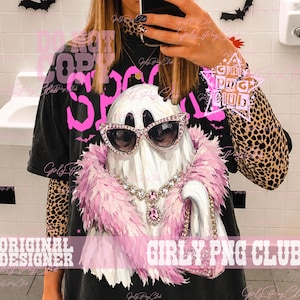 May include: A black t-shirt featuring a graphic of a white ghost wearing pink rhinestone cat-eye sunglasses, a pink feather boa, a silver-toned crystal necklace with a pink pendant, and holding a small pink glittery handbag.