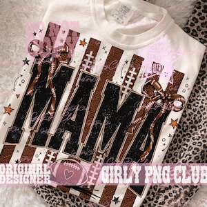 May include: A cream-colored t-shirt featuring a graphic design with the word MAMA in large black letters. The design includes brown football-textured stripes, football laces, brown bows, and small orange stars. The shirt has a crew neckline.