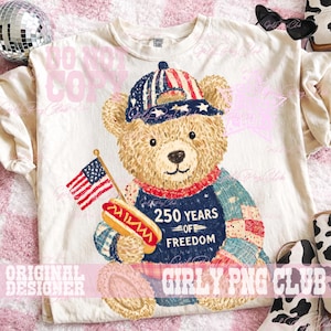 May include: Cream-colored t-shirt featuring a patriotic teddy bear design. The bear wears a hat and a vest with the text "250 YEARS OF FREEDOM" and holds an American flag and a hot dog. The design incorporates red, white, and blue colors.