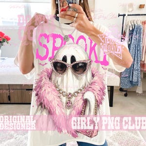 May include: A white t-shirt featuring a graphic of a ghost wearing pink rhinestone cat-eye sunglasses, a pink feather boa, a crystal necklace with a pink pendant, and holding a pink handbag. The word SPOOKY is printed in large pink letters.