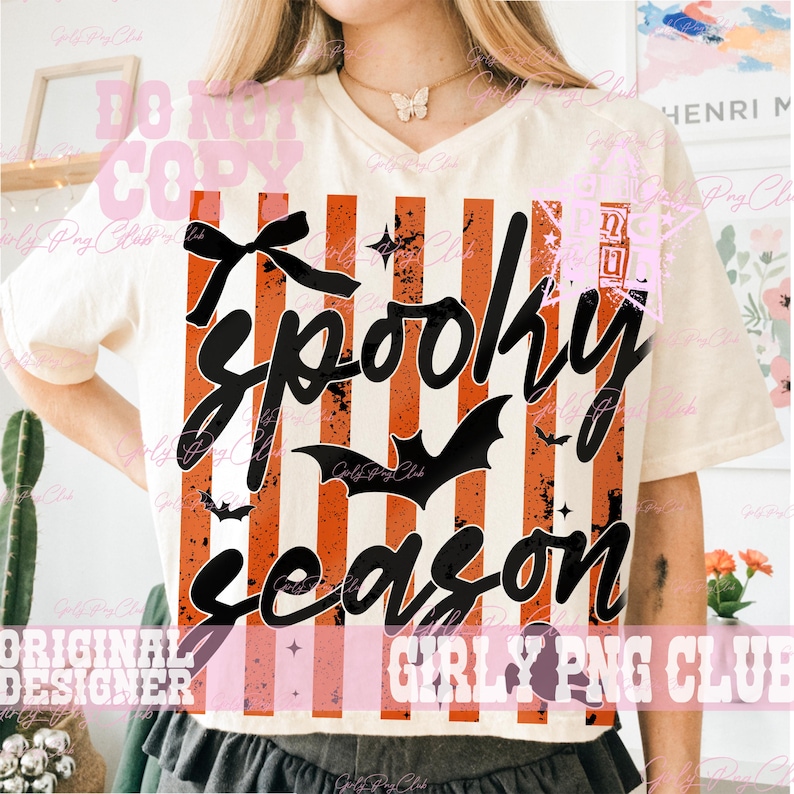 May include: A cream-colored short-sleeve t-shirt featuring a graphic print of vertical orange and white distressed stripes. The design includes black text that reads Spooky season, a black bow, and black bat silhouettes across the front.