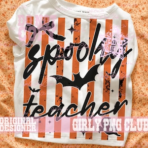 May include: A white t-shirt featuring vertical orange stripes and a distressed black graphic. The design includes the text "spooky teacher" in a cursive font, a large central bat, smaller bats, and a bow icon. The shirt has a round neckline.