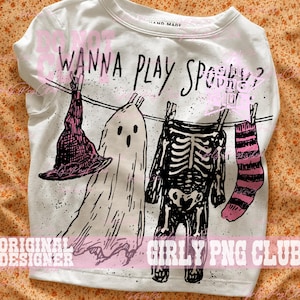 Op de afbeelding: Een wit T-shirt met ronde hals en een zwarte grafische print van een waslijn met een heksenhoed, een spook, een skelet en een gestreepte sok. De tekst WANNA PLAY SPOOKY? staat boven de afgebeelde items op het shirt gedrukt.