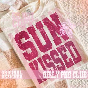 Pode incluir: Camiseta creme com gola redonda com as palavras "SUN KISSED" em letras de bloco rosa escuro desgastadas. A camiseta tem um visual vintage desbotado, com listras rosa verticais atr&aacute;s do texto. A camiseta est&aacute; dobrada em uma superf&iacute;cie estampada.