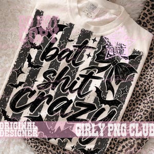 May include: A cream-colored t-shirt featuring a black graphic print with vertical stripes, bats, stars, and a large bow. The text reads "bat shit crazy" in a bold, brush-style font. The shirt has a crew neckline and a Comfort Colors brand label.