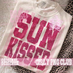 Pode incluir: Camiseta creme com as palavras "SUN KISSED" em letras rosa desbotadas. A camiseta tamb&eacute;m apresenta listras rosa verticais e o texto "ORIGINAL DESIGNER" e "GIRLY PNG CLUB". A camiseta &eacute; feita de um material macio.
