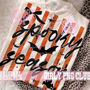 May include: A cream-colored t-shirt featuring a graphic print of vertical orange and white stripes with a distressed texture. The design includes the black text Spooky Season in a cursive font, accented by black bat silhouettes, a bow, and stars.