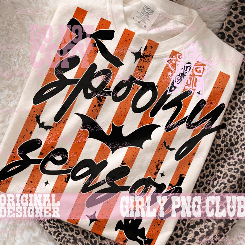 May include: A cream-colored t-shirt featuring a graphic print of vertical orange and white stripes with a distressed texture. The design includes the black text Spooky Season in a cursive font, accented by black bat silhouettes, a bow, and stars.