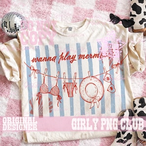 Op de afbeelding: Cr&egrave;mekleurig T-shirt met een rode en blauwe afbeelding. De afbeelding toont een waslijn met een bikini, ondergoed, een zonnehoed en harten. De tekst "wanna play mermaids" staat erboven. Het shirt heeft blauwe verticale strepen.