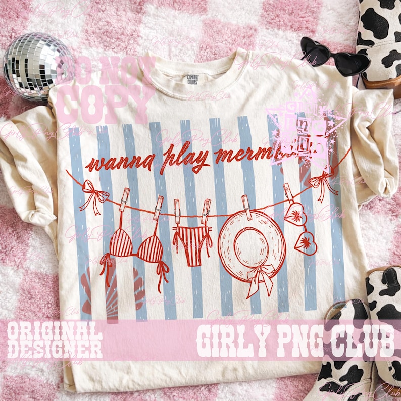 Op de afbeelding: Cr&egrave;mekleurig T-shirt met een rode en blauwe afbeelding. De afbeelding toont een waslijn met een bikini, ondergoed, een zonnehoed en harten. De tekst "wanna play mermaids" staat erboven. Het shirt heeft blauwe verticale strepen.