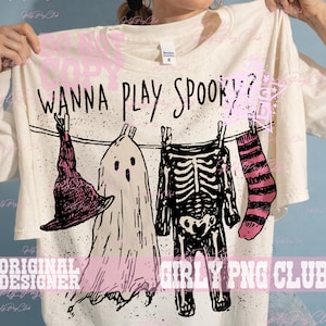 Op de afbeelding: Een off-white t-shirt met een zwarte afbeelding van een waslijn met een heksenhoed, een spook, een skeletkostuum en een gestreepte sok. De tekst "WANNA PLAY SPOOKY?" staat in het zwart boven de waslijn gedrukt met zwarte spikkels.