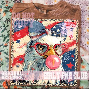 May include: Brown t-shirt featuring a graphic of an eagle wearing heart-shaped glasses and blowing a bubblegum bubble. The eagle is set against a backdrop of the American flag and fireworks. The text "Girly PNG Club" is visible.