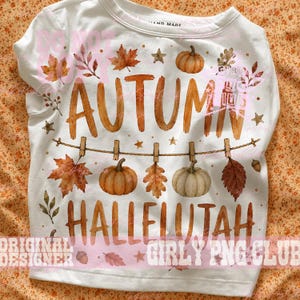 May include: A white crewneck t-shirt featuring a graphic print with the text AUTUMN HALLELUJAH in brown letters. The design includes watercolor-style illustrations of orange pumpkins, white pumpkins, brown autumn leaves, and small gold stars.