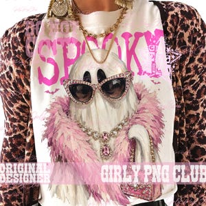 May include: A white t-shirt featuring a graphic of a ghost wearing pink cat-eye sunglasses, a pink feather boa, a jeweled necklace, and holding a pink handbag. The text "DO NOT BE SPOOKY" is printed in large pink letters above the ghost illustration.
