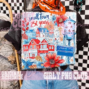 May include: A t-shirt with a vibrant graphic depicting a small town scene. The design includes red houses, blue details, and floral motifs. The text reads "small town 250 years". A water tower and street signs are also visible.