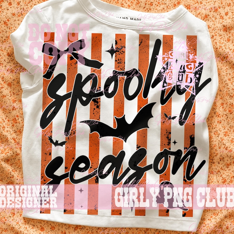 May include: A white t-shirt featuring a graphic print of vertical orange and black distressed stripes. The design includes the black text "spooky season" in a cursive font, a black bow, several black bat silhouettes, and small star icons.
