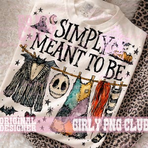 May include: A cream-colored t-shirt featuring the black text SIMPLY MEANT TO BE. The graphic includes a pinstriped suit, a skull, a patchwork dress, and long red hair hanging on a clothesline, accented with stars, bats, and pumpkins.
