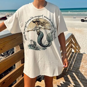 Puede incluir: Una camiseta blanca con un gr&aacute;fico de una sirena con un sombrero de vaquero y el texto "seaside cowgirl seaside cowgirl seaside cowgirl".