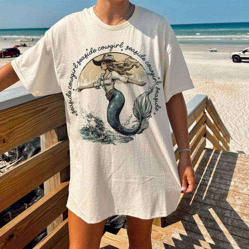 Puede incluir: Una camiseta blanca con un gr&aacute;fico de una sirena con un sombrero de vaquero y el texto "seaside cowgirl seaside cowgirl seaside cowgirl".