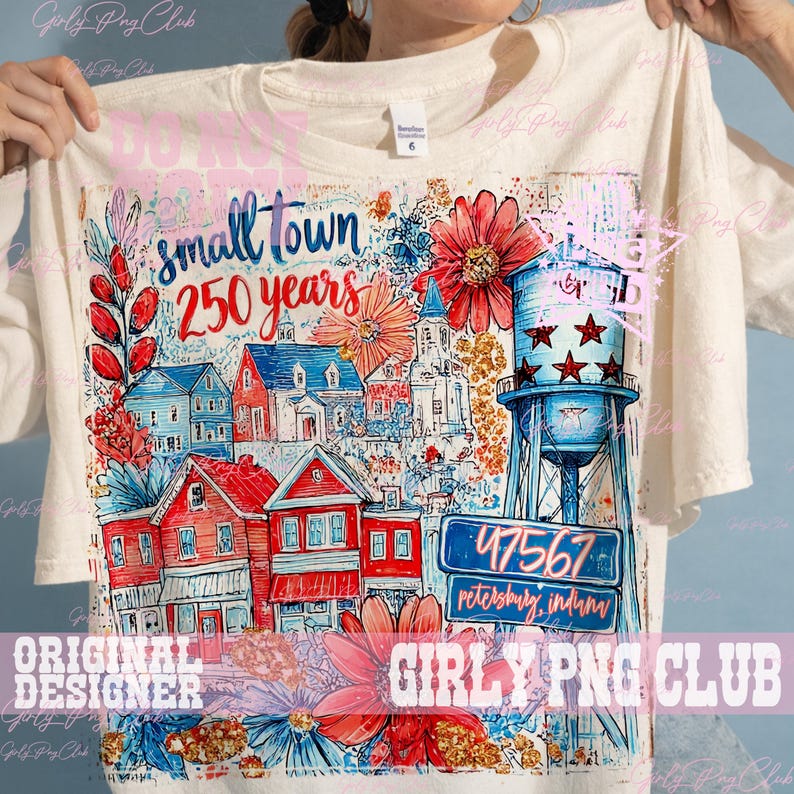 May include: Off-white t-shirt with a vibrant print depicting a small town scene. The design includes red and blue houses, a water tower, and the text "small town 250 years". The illustration also features floral elements and the town's postcode.
