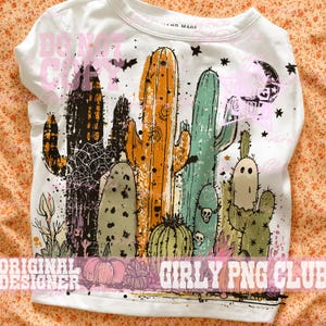 May include: A white t-shirt featuring a graphic print of various cacti in shades of orange, green, and black. The design includes small pumpkins, a crescent moon, stars, and a ghost-like figure. The text DO NOT COPY and GIRLY PNG CLUB is visible.